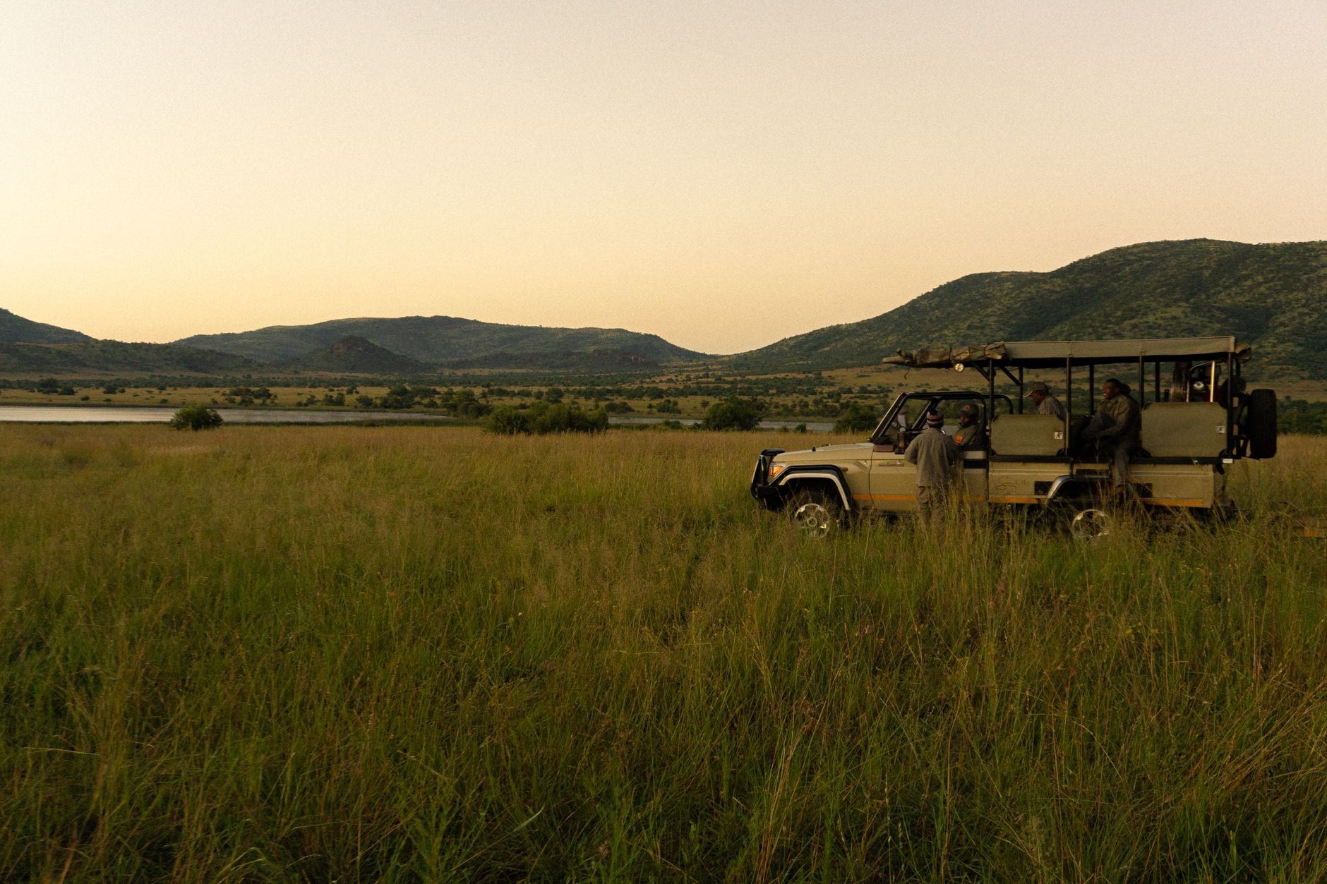 Ein Safarifahrzeug mit Besuchern parkt in offenem Grasland mit rollenden Hügeln und Wasser im Hintergrund im Pilanesberg-Nationalpark.