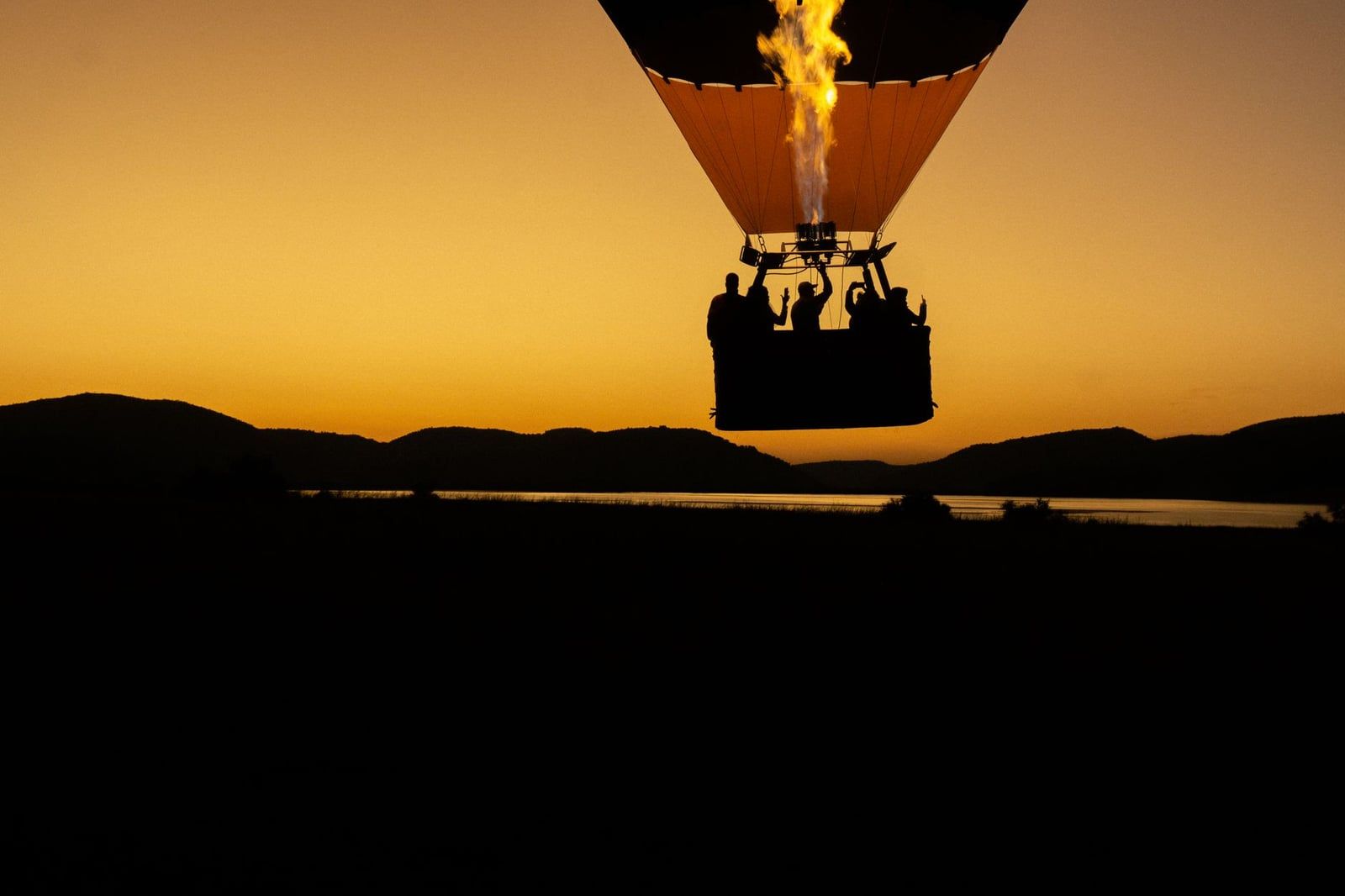 Silhouette of passengers in a hot air balloon basket with the burner firing against a golden sky above hills in Pilanesberg National Park.
